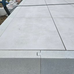 Niwińska Sarkiewicz Ogrody i Wnętrza - Nowoczesny taras z jasnoszarych płyt betonowych, połączonych na styk. Widoczne elementy mocujące i przygotowane miejsce na rośliny. Minimalistyczny design.