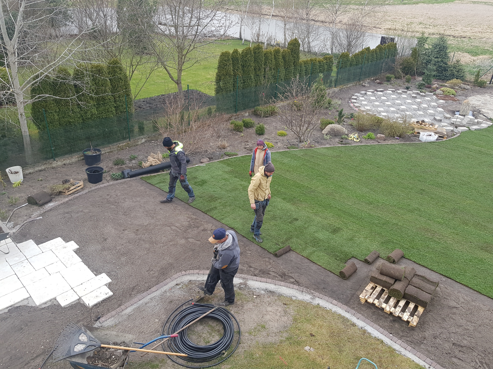 Montaż trawy z rolki w ogrodzie z widokiem z góry. Pracownicy rozkładają trawę, widoczne narzędzia i paleta z rolkami trawy. Częściowo ułożona kostka brukowa.
