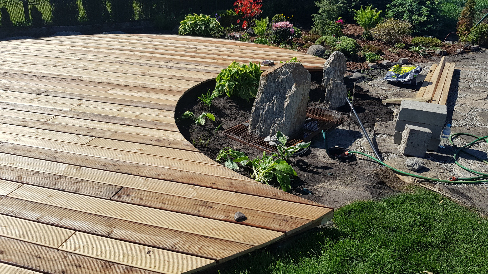 Drewniany taras z zaokrąglonym brzegiem, wkomponowany w ogród z kamieniami i roślinami. Widoczne elementy konstrukcyjne i narzędzia ogrodnicze.