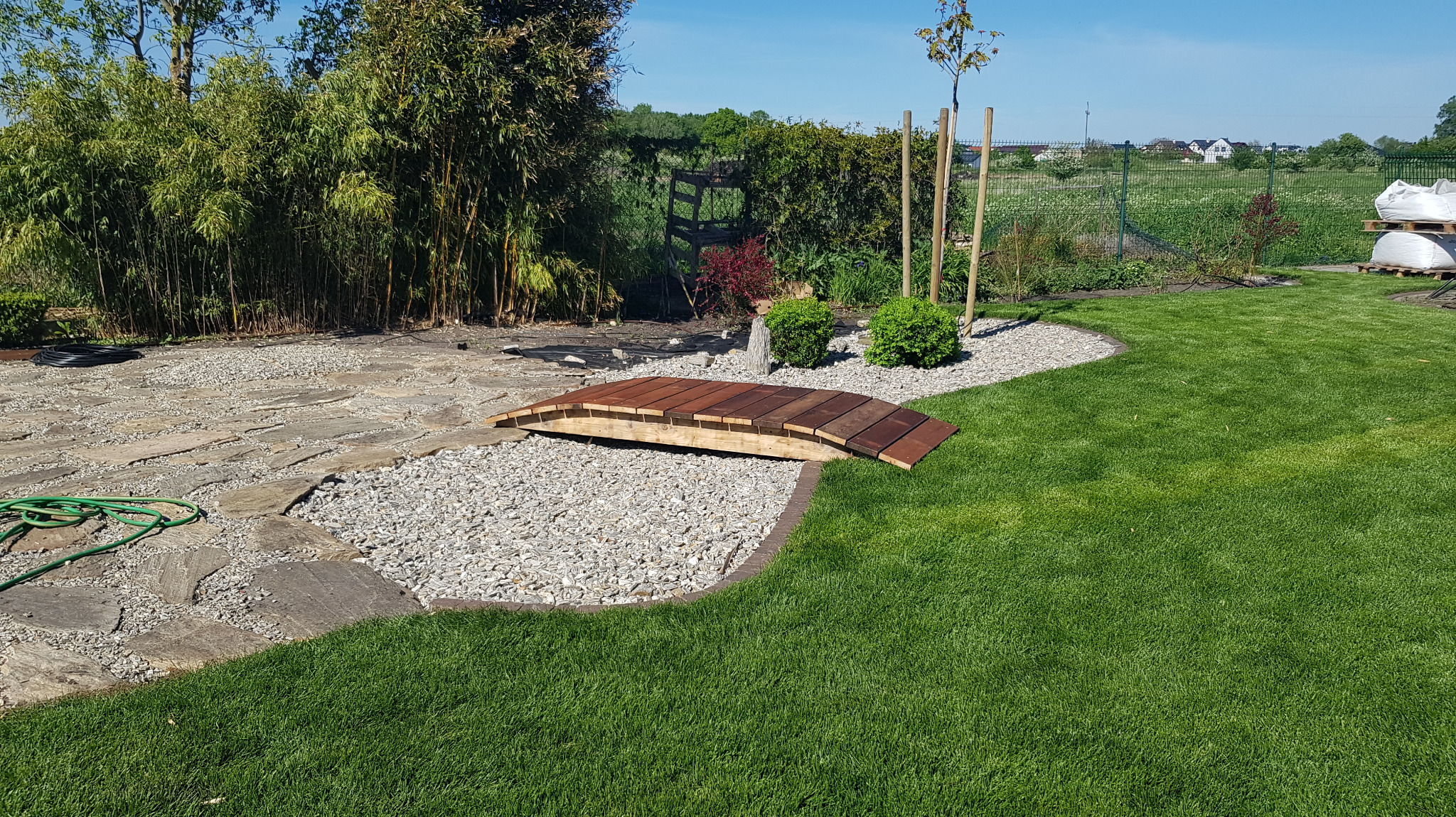 Ogród z drewnianym mostkiem nad kamiennym korytem, otoczony zielonym trawnikiem i bambusami. Elementy dekoracyjne i roślinność tworzą harmonijną całość.