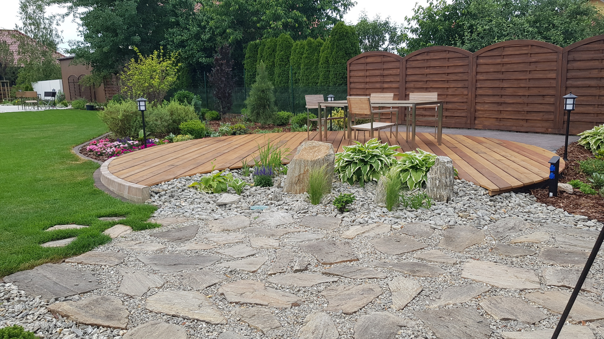 Ogród z drewnianym tarasem, kamienną ścieżką i ozdobnymi roślinami. Widoczne oświetlenie i drewniane ogrodzenie w tle. Uwagę przykuwa okrągły kształt tarasu.