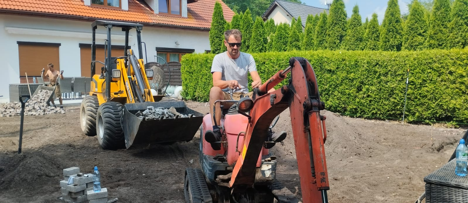 Pracownik w okularach na mini koparce, obok ładowarka z kamieniami. Przygotowanie terenu pod kostkę brukową na tle domu i zielonych tuj.