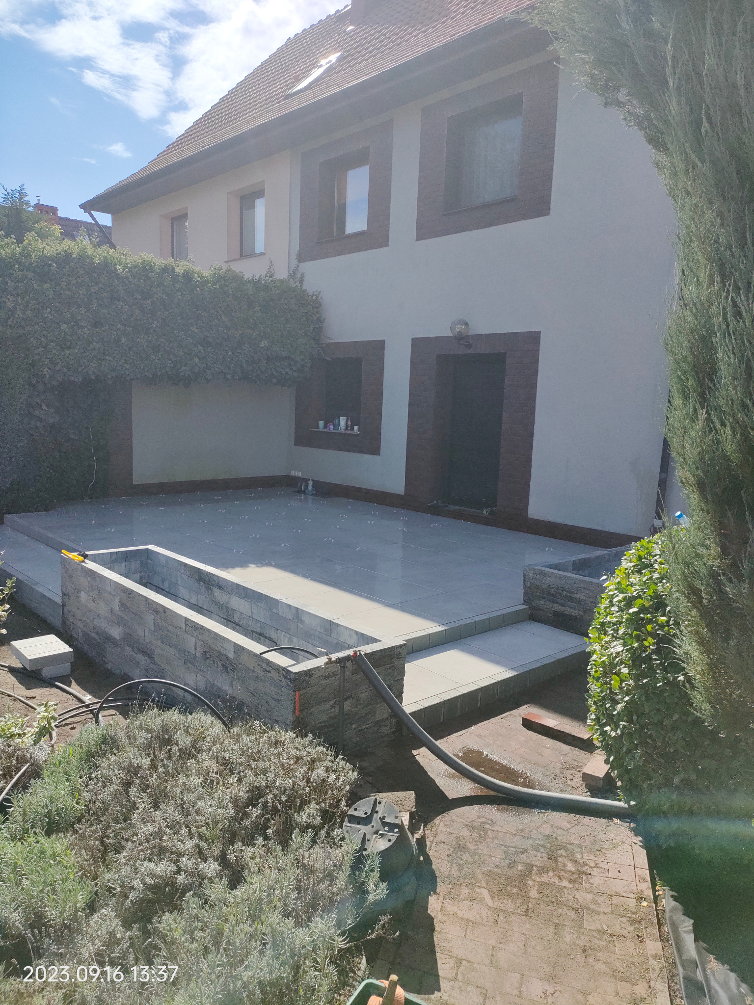 Nowoczesny taras z szarych płyt, obudowany podwyższoną rabatą z kamienia. Widoczne elementy systemu nawadniania i fragment ogrodu. Jasne, naturalne światło.