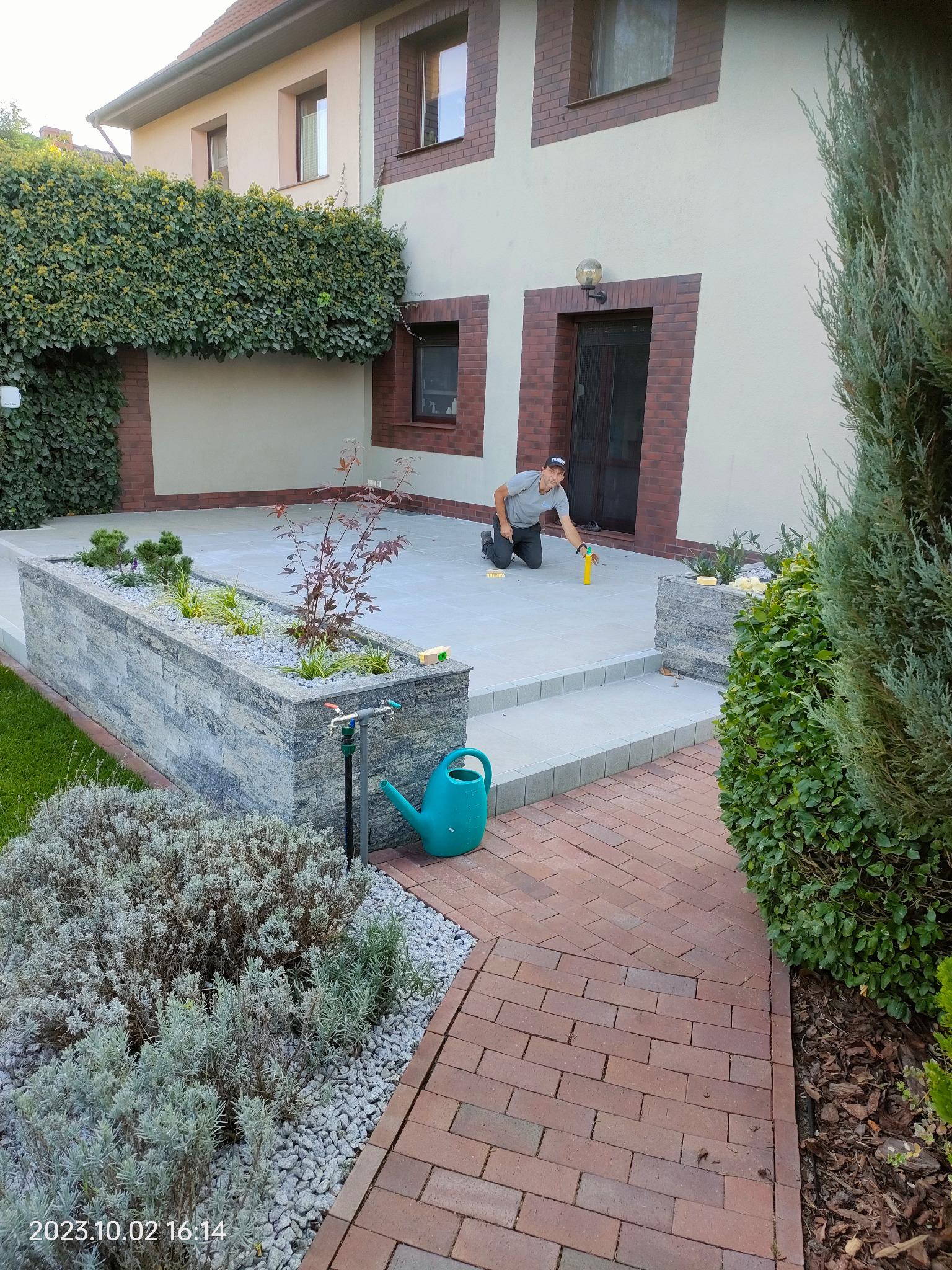 Ukończony taras z kostki, z roślinami w donicach. Mężczyzna czyści fugi. Widoczny kranik i konewka. Czerwona kostka brukowa ścieżki. Dom z ceglanymi elementami.