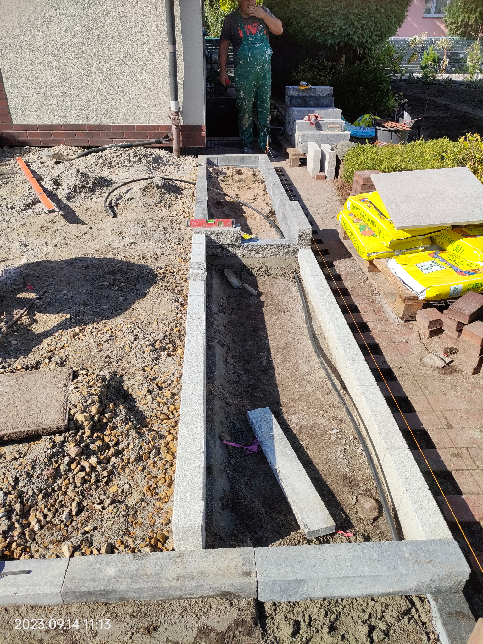 Budowa obrzeży rabatki z betonowych elementów. Widoczny fragment ogrodu w trakcie prac ziemnych, narzędzia i materiały budowlane. Prace ogrodnicze w słoneczny dzień.