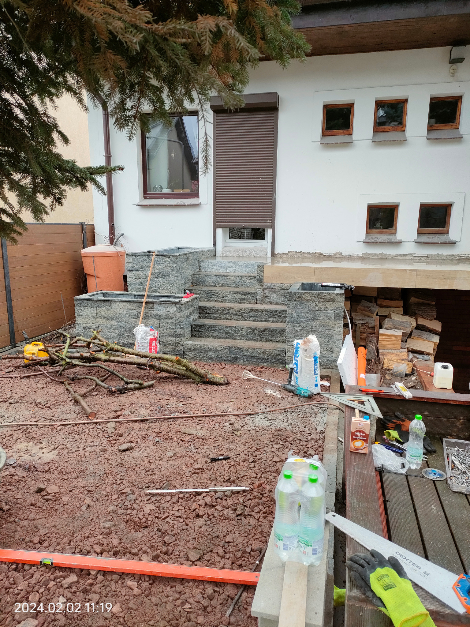 Budowa schodów wejściowych z kamienia, widoczne narzędzia i materiały budowlane. Etap prac wykończeniowych, przygotowanie terenu pod dalsze prace.