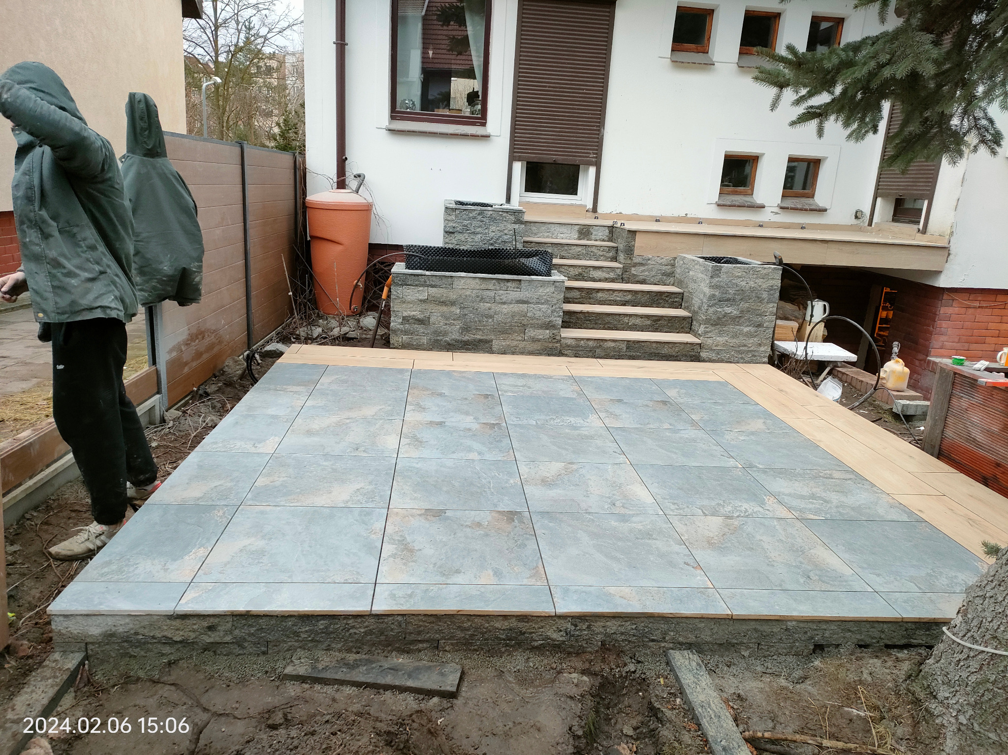 Tarasy z płytek i schody z kamienia na zewnątrz domu. Widoczny fragment ogrodu i osoba w kurtce. Ujęcie z dołu, podkreślające detale wykończenia.