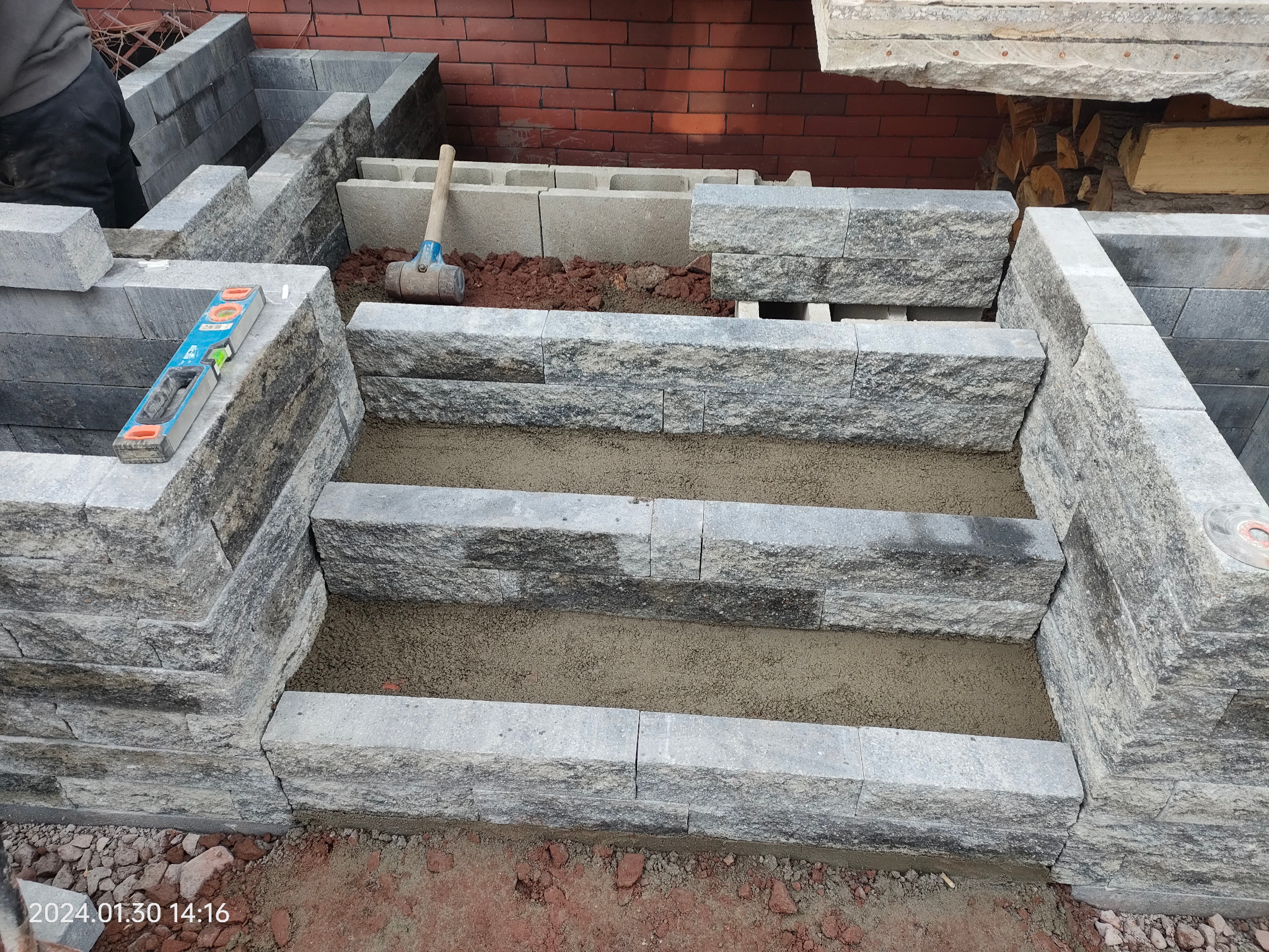 Budowa schodów zewnętrznych z szarych bloków kamiennych. Poziomica na murku, gumowy młotek obok. Widoczny etap prac, podsypka piaskowa na stopniach.