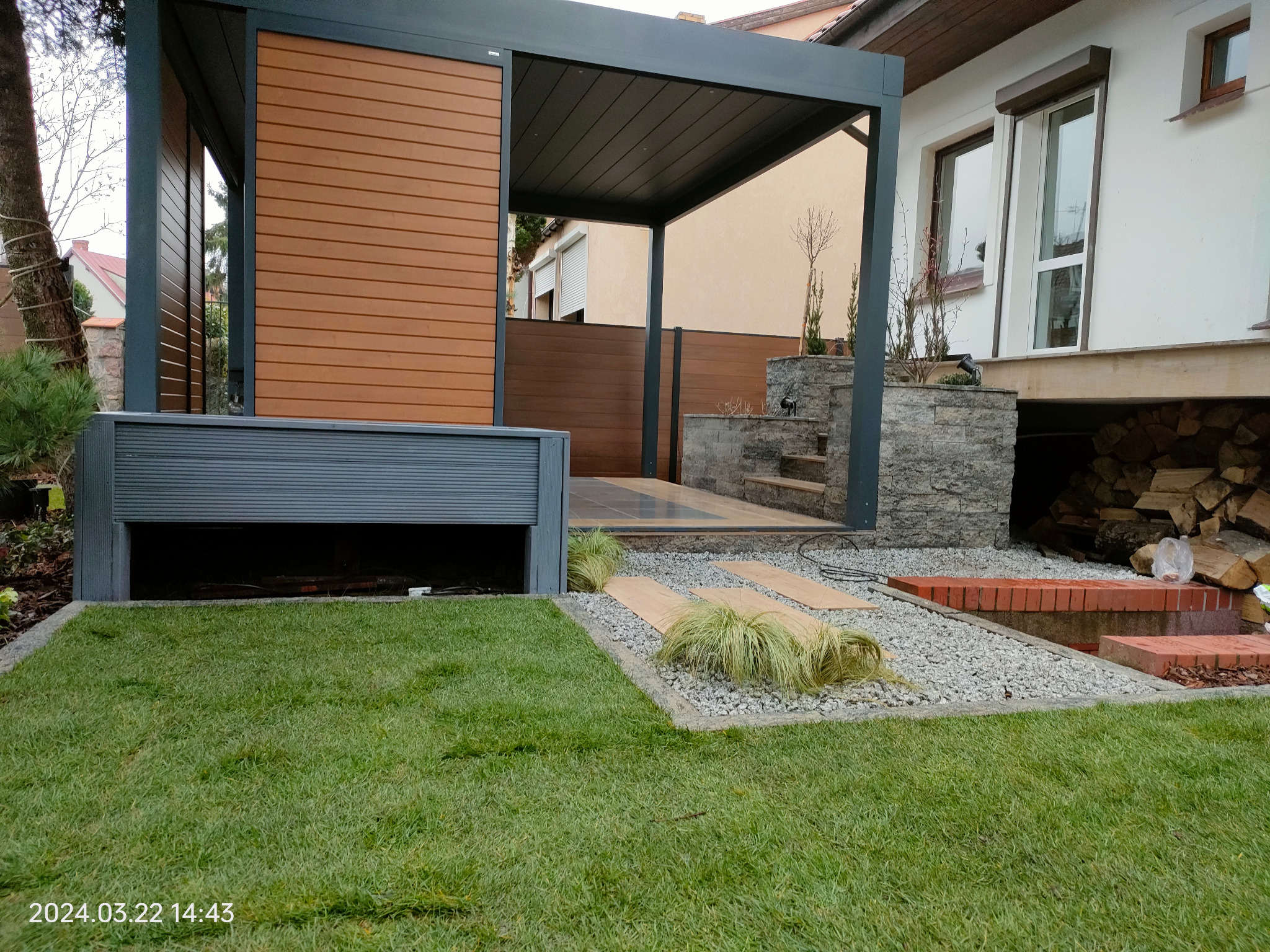 Nowoczesna pergola z drewnianą zabudową i roletą, otoczona świeżą trawą i kamienną ścieżką. Elementy małej architektury ogrodowej wkomponowane w otoczenie domu.