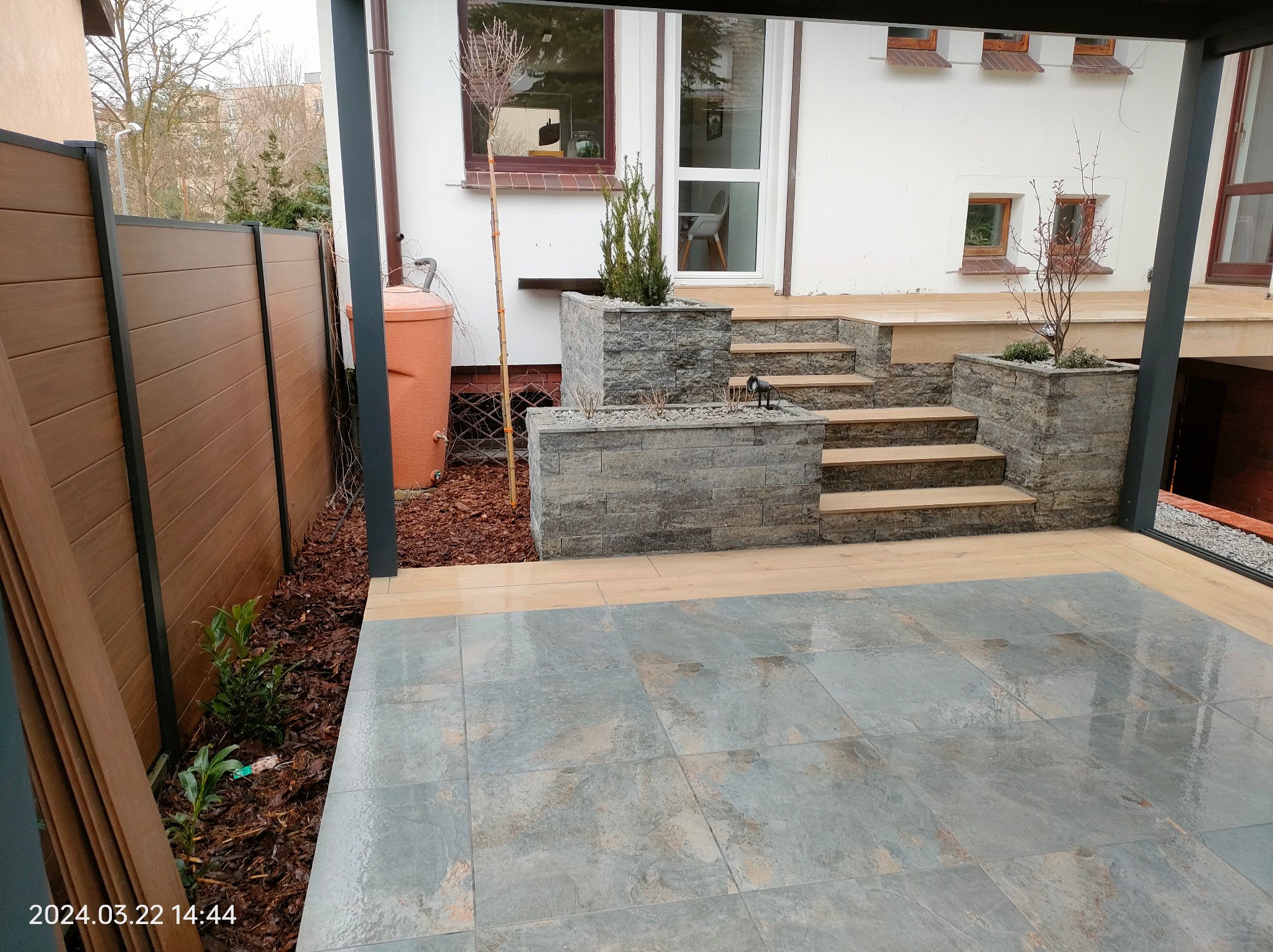 Nowoczesny taras z kamiennymi donicami i drewnianymi schodami, otoczony brązowym płotem. Widoczny zbiornik na deszczówkę i fragment budynku z oknami.