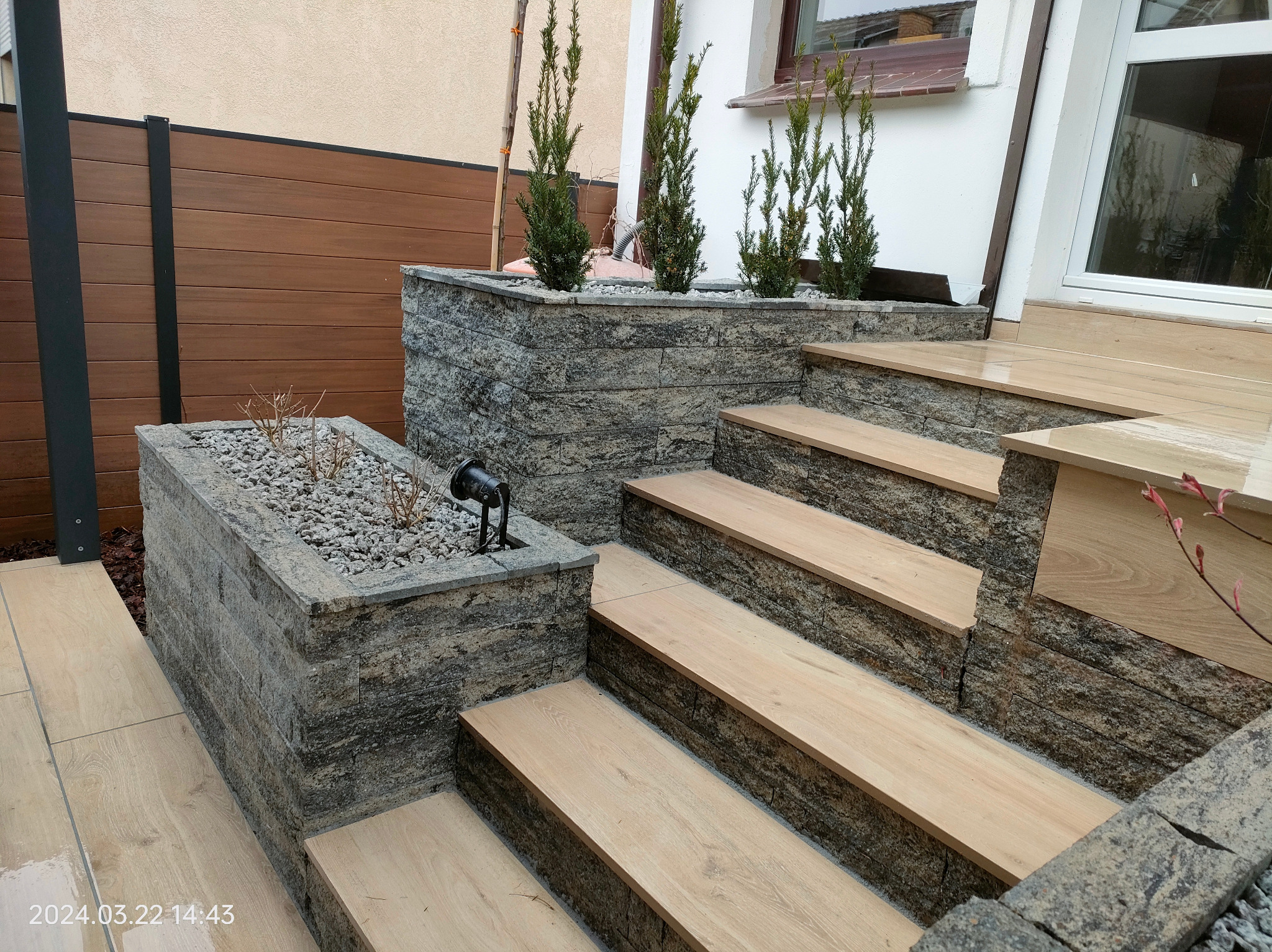 Kamienne donice z roślinami i schody z drewnopodobnych płyt tworzą nowoczesny ogród. Widoczne oświetlenie punktowe i drewniane panele ogrodzeniowe.