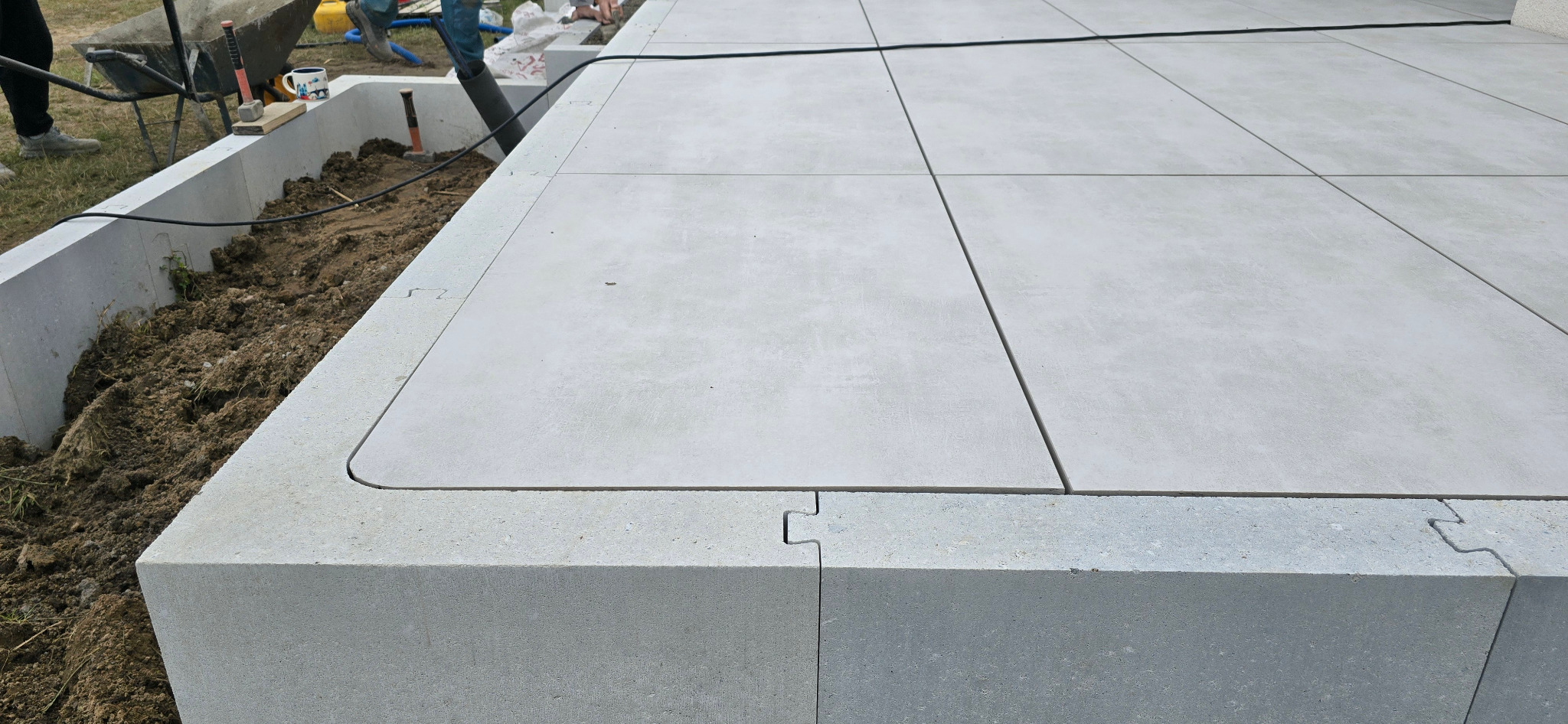 Nowoczesny taras z jasnoszarych płyt betonowych, połączonych na styk. Widoczne elementy mocujące i przygotowane miejsce na rośliny. Minimalistyczny design.