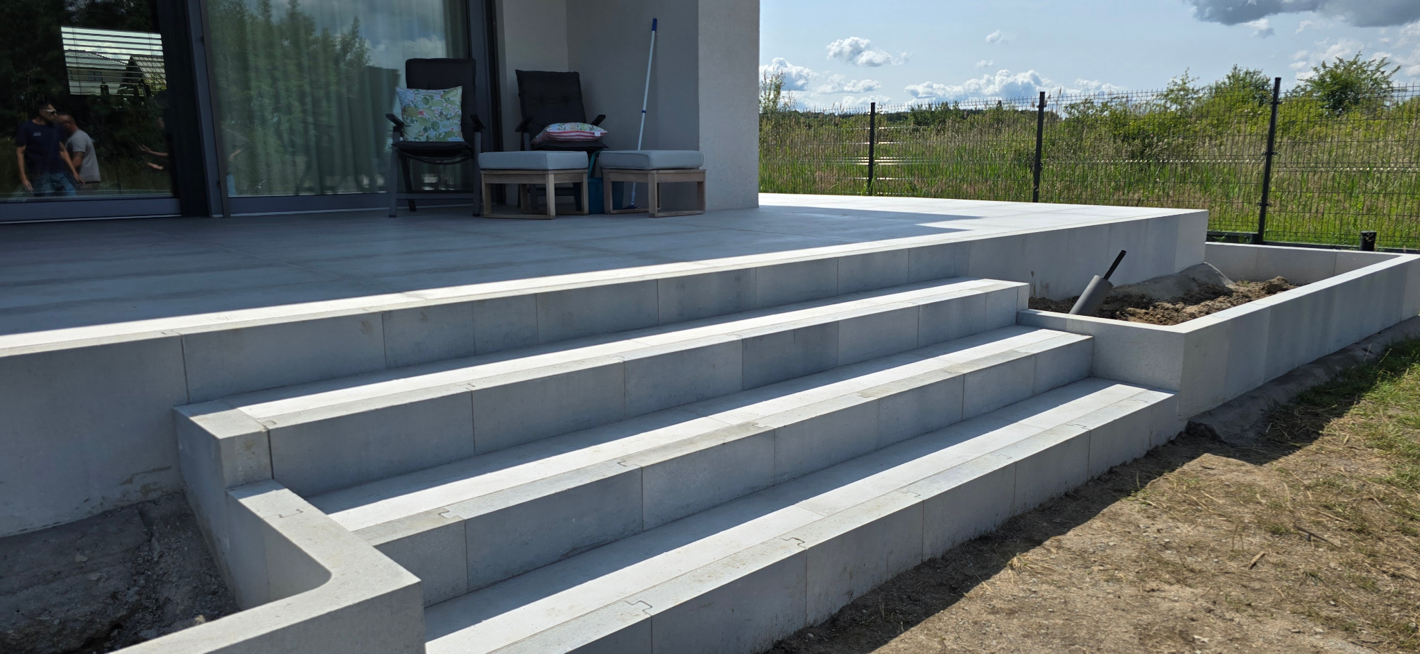 Nowoczesne schody wejściowe z jasnoszarej kostki brukowej prowadzące na taras z meblami ogrodowymi i rabatą. Widok na zewnątrz przez szklane drzwi.