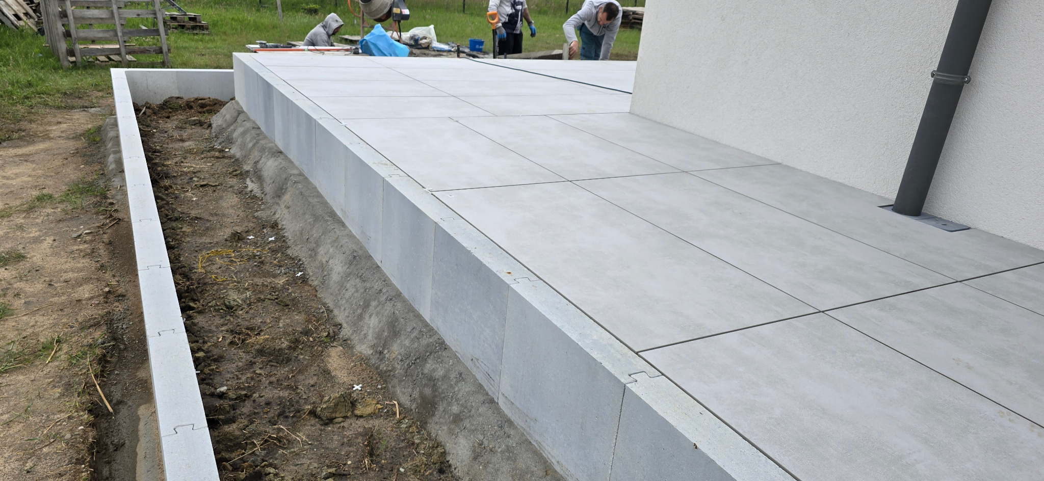 Jasnoszara, kwadratowa kostka brukowa ułożona na tarasie przydomowym. Widoczny fragment ściany budynku i rynna. W tle pracownicy podczas układania.