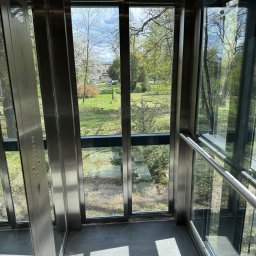 POWERLIFT Sp. z o.o. - Wnętrze nowoczesnej windy z panoramicznym widokiem na park. Stalowe wykończenia, szklane ściany, wpadające promienie słoneczne. Widok na zieleń i budynek w tle.