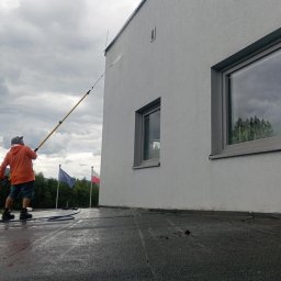 RKubica mycie cisnieniowe - Pracownik w pomarańczowej kurtce czyści elewację budynku za pomocą lancy teleskopowej. W tle widoczne flagi Polski i Unii Europejskiej.