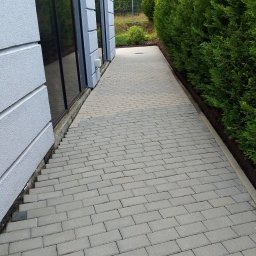 RKubica mycie cisnieniowe - Ścieżka z szarej kostki brukowej, biegnąca wzdłuż budynku z dużymi oknami i żywopłotu. Widok z góry, ukazujący regularny wzór ułożenia kostki.