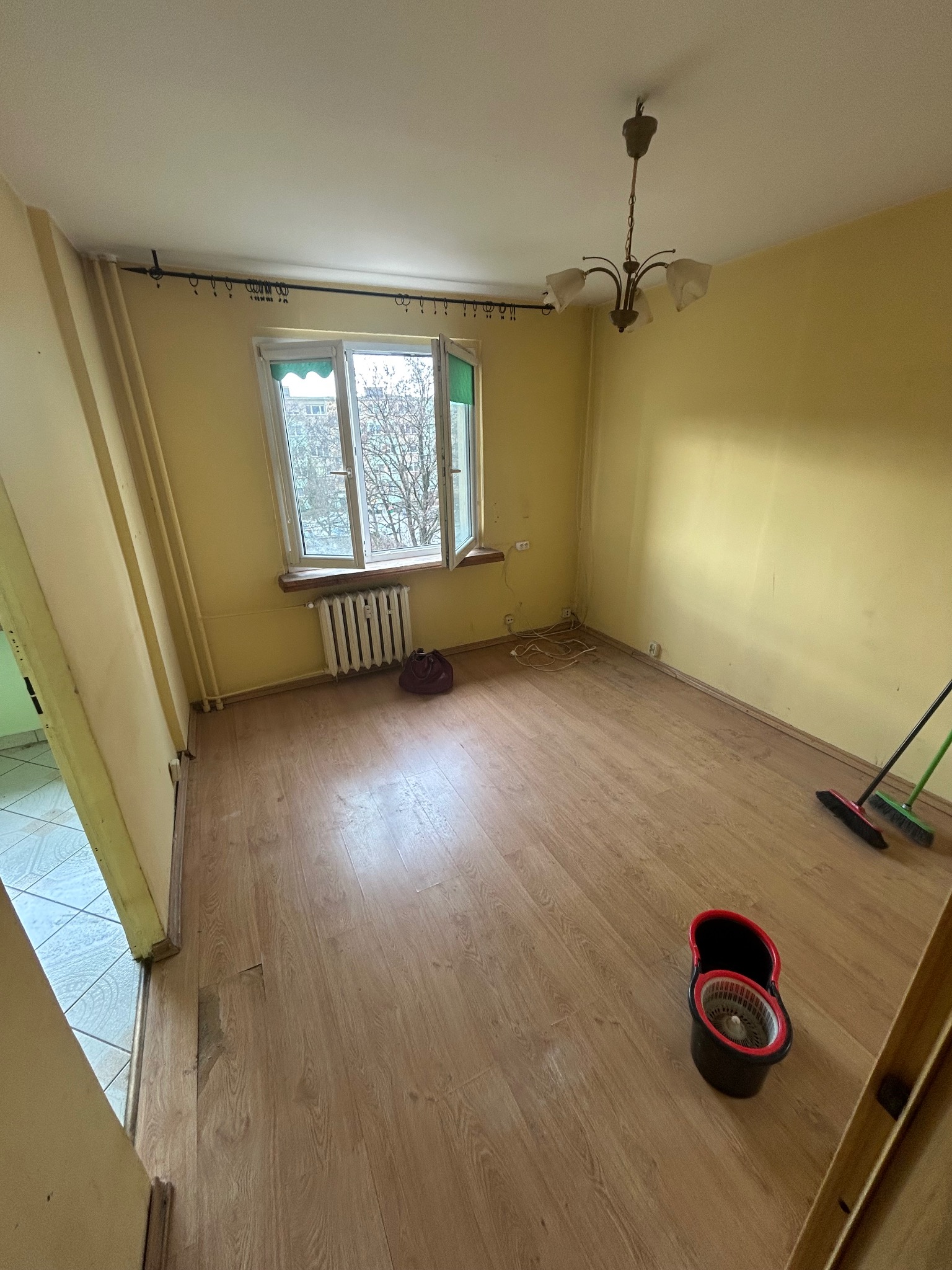 Pusty pokój z żółtymi ścianami i drewnianą podłogą, mop i wiadro w rogu, otwarte okno z widokiem na drzewa. Przygotowanie do sprzątania.