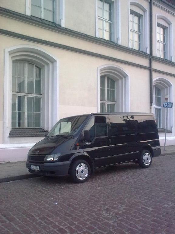 Czarny bus Ford Transit zaparkowany na brukowanej ulicy przed jasnym budynkiem z dużymi oknami, obok widoczny znak TAXI.