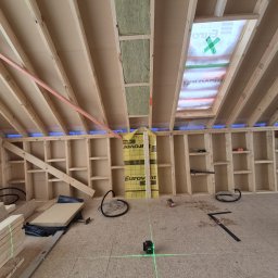 PK Konstrukcje i Wykończenia - Poddasze w trakcie adaptacji: widoczne drewniane elementy konstrukcyjne, izolacja, okno dachowe zabezpieczone folią i laser krzyżowy wyznaczający poziom.