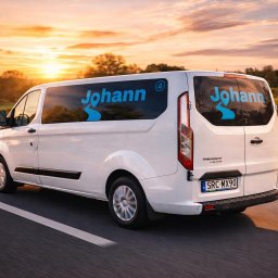 Johann - Biały bus marki Ford Transit z niebieskim logo 'Johann' na tle zachodzącego słońca, jadący po asfalcie. Widok z tyłu i boku, dynamiczne ujęcie w ruchu.