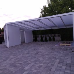 FSR SERHII REUTSKYI - Nowoczesna, biała pergola metalowa z przezroczystym zadaszeniem na posesji z kostki brukowej. Widoczny fragment domu i zieleń w tle. Osoba w oddali po lewej.