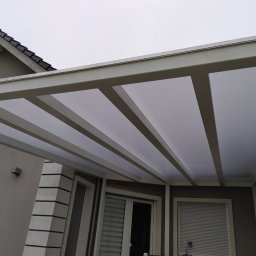 FSR SERHII REUTSKYI - Nowoczesna pergola z białymi belkami i przezroczystym zadaszeniem, przylegająca do szarego budynku z oknami i roletami. Widok z dołu, pochmurne niebo w tle.