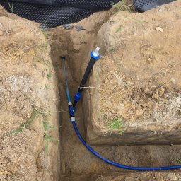 Usługi Hydrauliczne Wojciech Antosz - Niebieska rura wodna w wykopie ziemnym, z czarną folią fundamentową w tle. Widoczne złącza i fragment instalacji. Prace ziemne, instalacja wodociągowa.