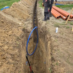 Usługi Hydrauliczne Wojciech Antosz - Wykop z niebieską rurą w środku, pomarańczowe rury na górze, dwóch pracowników w tle. Prace ziemne pod instalacje wod-kan na działce.