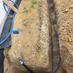 Usługi Hydrauliczne Wojciech Antosz - Wykop z ułożoną niebieską rurą instalacyjną, widoczne złączki i fragment ściany fundamentowej z izolacją. Prace ziemne w toku, widoczna łopata i narzędzia.