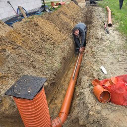 Usługi Hydrauliczne Wojciech Antosz - Montaż rur kanalizacyjnych w wykopie, pracownik sprawdza poziom, w tle koparka i budynek w trakcie budowy. Pomarańczowe rury z tworzywa sztucznego.