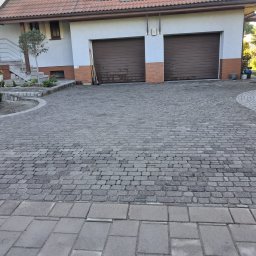 M&R Spółka z ograniczoną odpowiedzialnością - Podjazd i ścieżki z szarych kamieni przed domem z dwoma brązowymi bramami garażowymi. Nawierzchnia łączy prostokątne płyty z mniejszymi, nieregularnymi elementami, tworząc zakrzywione rabaty.