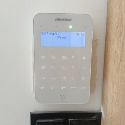 Primeron Sp. z o.o. - Panel alarmowy Hikvision z wyświetlaczem LCD, zamontowany na ścianie w Krakowie. Widoczny czarny włącznik światła pod panelem. Alarm domowy.