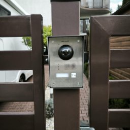 Primeron Sp. z o.o. - Zamontowany wideodomofon Hikvision na brązowej bramie posesji. Widoczny fragment chodnika z kostki i budynek w tle. Ujęcie z bliska, skupione na urządzeniu.