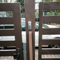 Primeron Sp. z o.o. - Zamontowany wideodomofon z przyciskiem na brązowej bramie posesji, widok z bliska. W tle zaparkowany biały samochód i budynek mieszkalny.