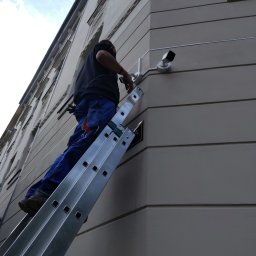 Primeron Sp. z o.o. - Instalator na drabinie montuje kamerę monitoringu na rogu kamienicy. Widok z dołu, błękitne niebo w tle. Dwie kamery już zamontowane.