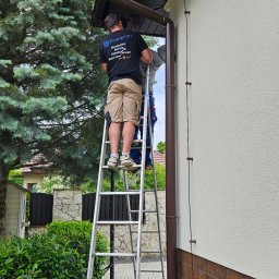 Primeron Sp. z o.o. - Dwóch monterów na drabinie instaluje przewody na elewacji domu, prawdopodobnie system monitoringu. Widoczne elementy instalacji i narzędzia.