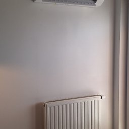 Primeron Sp. z o.o. - Biały klimatyzator Haier zamontowany na ścianie nad grzejnikiem, widoczny fragment okna z zasłoną. Minimalistyczne wnętrze.