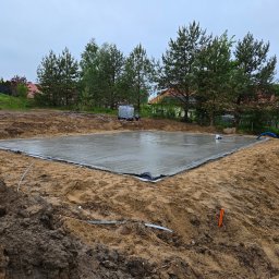 KOPTECH Sebastian Konopka - Wylana płyta fundamentowa na gruncie w Nikielkowie, zabezpieczona folią. Widoczne przygotowane przyłącza i otaczający teren budowy z drzewami w tle.
