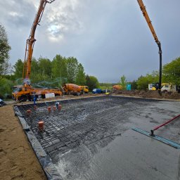 KOPTECH Sebastian Konopka - Wylewanie betonu na zbrojenie fundamentów za pomocą pompy do betonu. Widoczne pomarańczowe rury i ekipa budowlana. W tle kilka betoniarek i pochmurne niebo.