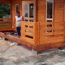 KONKRET - Budowa drewnianego domku letniskowego z tarasem w Gdańsku. Widoczny pracownik podczas montażu balustrady. Konstrukcja osadzona na betonowych bloczkach.