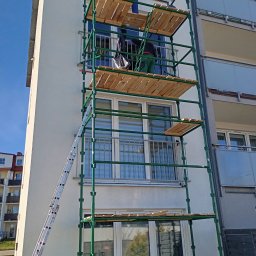 KONKRET - Dwóch pracowników na zielonym rusztowaniu przy białym budynku wielorodzinnym. Jeden w pomarańczowej bluzie, drugi w zielonych spodniach. Widoczna drabina i błękitne niebo.