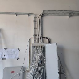 Marek Warumzer - Rozdzielnia elektryczna z licznymi kablami w białej osłonie, prowadzącymi do metalowych korytek na ścianie. Widoczny schemat instalacji i falownik Fronius.