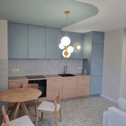 myWall - Jasna kuchnia po remoncie: drewniane szafki, okrągły stół i designerska lampa. Ściany w odcieniach szarości i błękitu, mozaika na podłodze.