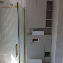 Cw Budownictwo - Nowoczesna łazienka z kabiną prysznicową ze złotymi detalami, podwieszaną toaletą i szafkami w odcieniach szarości. Minimalistyczny design i pionowe żłobienia na ścianie.