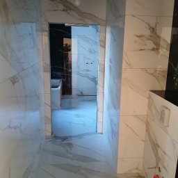 Cw Budownictwo - Wykończona łazienka z marmurowymi płytkami na podłodze i ścianach. Widoczny fragment sufitu podwieszanego i instalacja sanitarna. W głębi widoczne wyposażenie łazienki.
