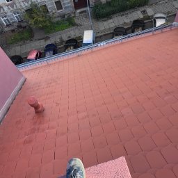 Firma Usługowa Kacper Buczek - Czerwony dach z dachówki ceramicznej widziany z góry, z fragmentem buta osoby stojącej na dachu. W tle budynki mieszkalne i zaparkowane samochody.