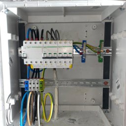 Volt-Tech - Wnętrze skrzynki elektrycznej z zamontowanymi bezpiecznikami i przewodami. Widoczne złącza, listwy DIN i kable w różnych kolorach. Instalacja w białej obudowie.