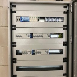 Volt-Tech - Otwarta skrzynka elektryczna z zainstalowanymi bezpiecznikami i przewodami, widoczne czarne rurki instalacyjne przy suficie, ściana z szarego betonu komórkowego.
