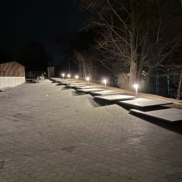 Volt-Tech - Oświetlona ścieżka z płyt i lamp wzdłuż rzeki nocą. Widoczne drzewa bez liści i odbicia świateł w wodzie. Geometryczny wzór kostki brukowej.